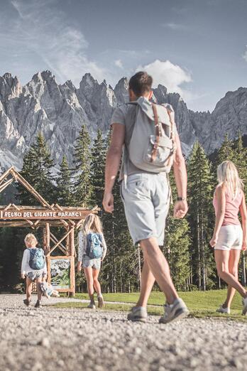 Zwei Kinder spielen auf einem Spielplatz vor einer Berglandschaft; im Vordergrund geht eine Frau über einen Kiesweg, im Hintergrund sind Wiese, Wald und hohe Felsgipfel zu sehen.