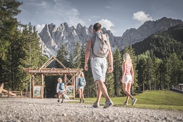 Zwei Kinder spielen auf einem Spielplatz vor einer Berglandschaft; im Vordergrund geht eine Frau über einen Kiesweg, im Hintergrund sind Wiese, Wald und hohe Felsgipfel zu sehen.