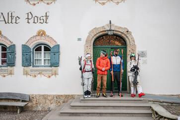 Vier Personen in Skiausrüstung stehen mit Skiern vor einem historischen Gebäude