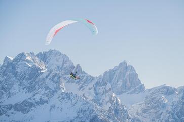 Ein Paragleiter schwebt über eine weitläufige Berglandschaft mit Schneebedeckten Gipfeln.
