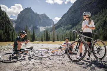 Zwei Personen mit Fahrradhelmen stehen mit ihren Fahrrädern auf einem steinigen Aussichtspunkt in einem Bergtal; im Hintergrund sind Wald, hohe Felsgipfel und ein blauer Himmel mit Wolken zu sehen.
