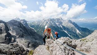 Zwei Kletterer steigen einen Felsen hoch; im Hintergrund sind Gebirge, Täler und der blaue Himmel mit weißen Wolken zu sehen.