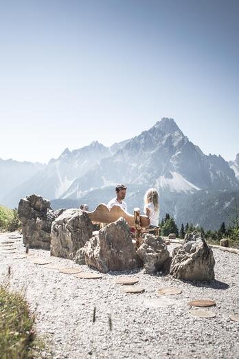 Ein Mann und ein Kind sitzen auf einer Holzbank; im Hintergrund ist eine massive Berglandschaft zu sehen.
