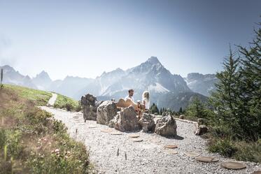 Ein Mann und ein Kind sitzen auf einer Holzbank; im Hintergrund ist eine massive Berglandschaft zu sehen.