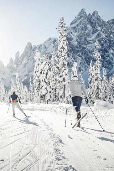Zwei Langläufer fahren auf einer verschneiten Loipe durch eine sonnige Winterlandschaft mit schneebedeckten Bäumen und Bergen im Hintergrund.