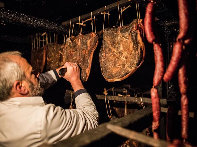 Ein Mann schneidet in einem dunklen Reifekeller hängende Speck- und Wurstwaren mit einem Messer.