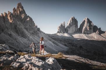 Ein Mann und eine Frau wandern in den Dolomiten, im Hintergrund erheben sich die Drei Zinnen