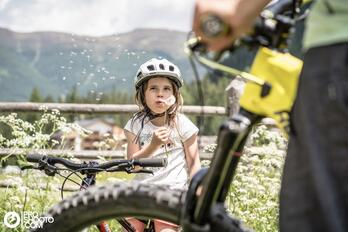Ein Mädchen mit Fahrradhelm steht neben einem Fahrrad; im Vordergrund ist ein weiteres Fahrrad unscharf zu sehen, im Hintergrund eine Steinmauer und Berge.
