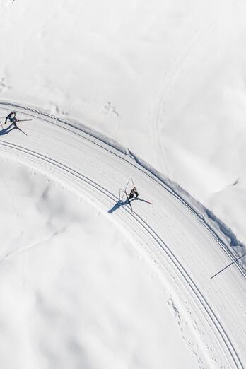 Luftaufnahme einer verschneiten Kurve mit Spuren im Schnee und zwei Personen auf einer Winterstrecke.