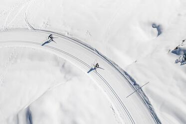 Luftaufnahme einer verschneiten Kurve mit Spuren im Schnee und zwei Personen auf einer Winterstrecke.