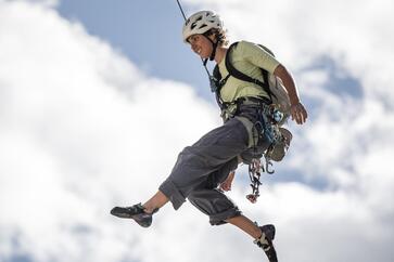 Kletterer mit Helm und Rucksack hängt gesichert an einem Seil vor blauem Himmel mit weißen Wolken.