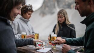 Mehrere Personen sitzen gemeinsam an einem Tisch in den Sextner Dolomiten, trinken Bier und spielen Karten