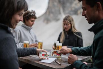 Mehrere Personen sitzen gemeinsam an einem Tisch in den Sextner Dolomiten, trinken Bier und spielen Karten