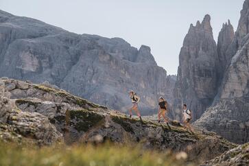 Drei Wanderer in weitläufigem Gebirge; im Hintergrund ist massive Berglandschaft zu sehen.