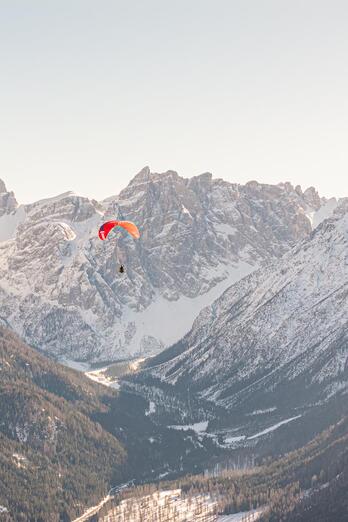 Zwei Paragleiter schwebt über einer weitläufigen Berglandschaft mit schneebedeckten Gipfeln im Hintergrund.