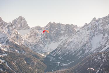 Zwei Paragleiter schwebt über einer weitläufigen Berglandschaft mit schneebedeckten Gipfeln im Hintergrund.