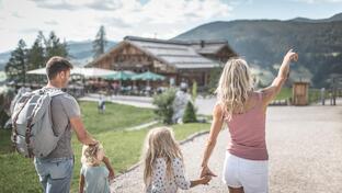 Eine Familie geht über einen weitläufigen Platz in Richtung eines Gebäudes mit Spielbereich; im Hintergrund sind Bäume, Berge und ein bewölkter Himmel zu sehen.