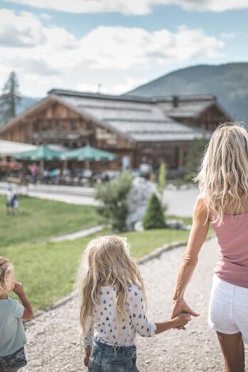Eine Familie geht über einen weitläufigen Platz in Richtung eines Gebäudes mit Spielbereich; im Hintergrund sind Bäume, Berge und ein bewölkter Himmel zu sehen.