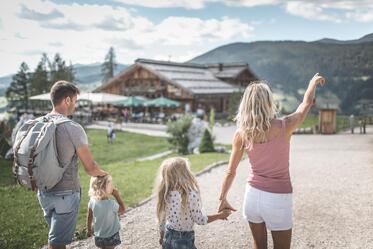 Eine Familie geht über einen weitläufigen Platz in Richtung eines Gebäudes mit Spielbereich; im Hintergrund sind Bäume, Berge und ein bewölkter Himmel zu sehen.