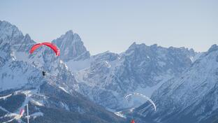 Zwei Paragleiter fliegen vor einer weitläufigen Berglandschaft mit schneebedeckten Gipfeln.