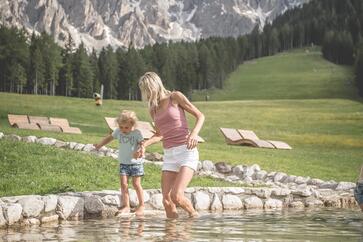 Eine Frau und ein Kind stehen barfuß in einem flachen Bach vor einer Berglandschaft; im Hintergrund sind eine Wiese, Wald und markante Felsgipfel zu sehen.