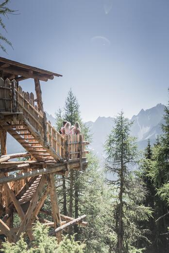 Ein Mann, eine Frau und zwei Kinder sitzen auf einem Holzgerüst im Wald und blicken in Richtung einer Berglandschaft.